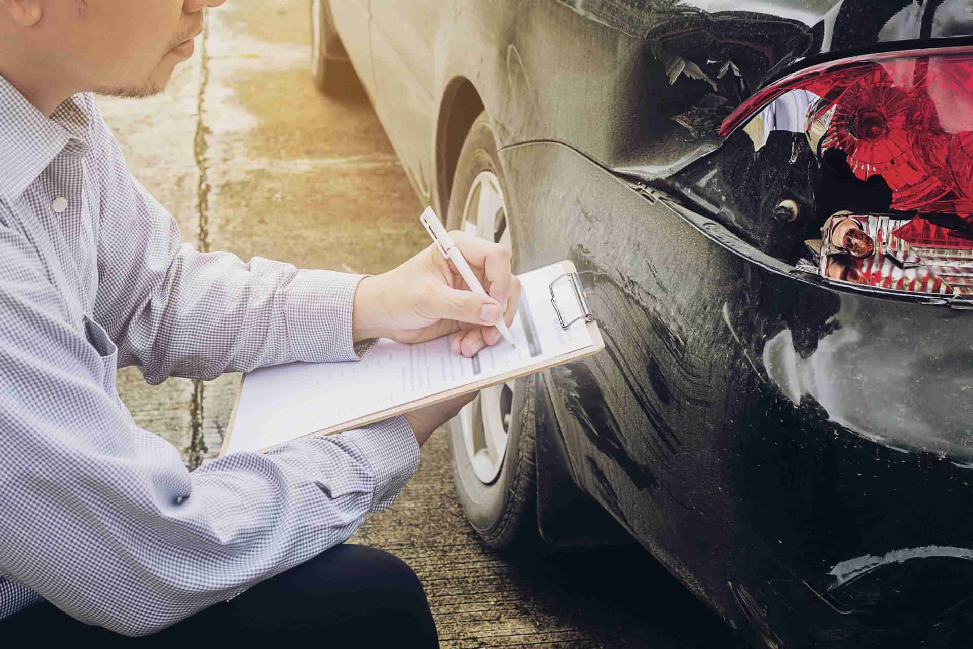 Person with a clipboard examines the damaged rear bumper of a black car on a sunny day.
