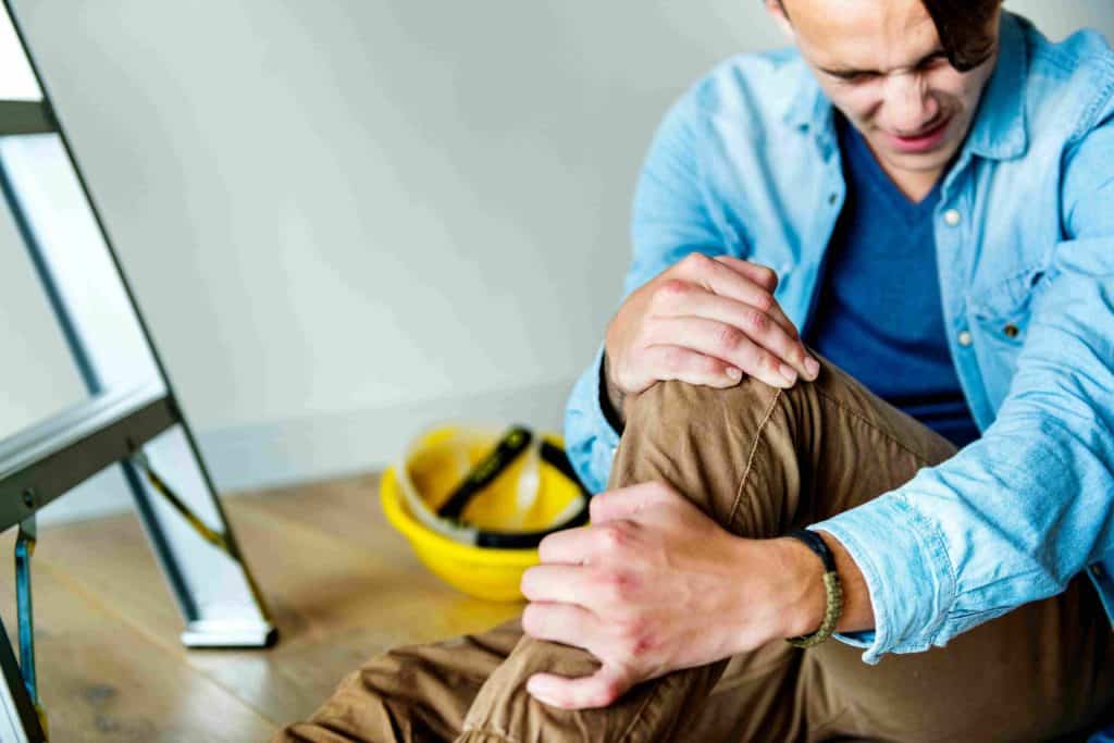 A man in a blue shirt sits on the floor, holding his knee in pain. A yellow hard hat and a ladder are nearby.