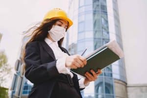 Woman in a suit and hard hat, wearing a mask, writes in a notebook outside a modern glass building.