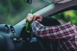 Person in a plaid shirt leans on a car steering wheel, holding a green bottle, with rain visible on the window.