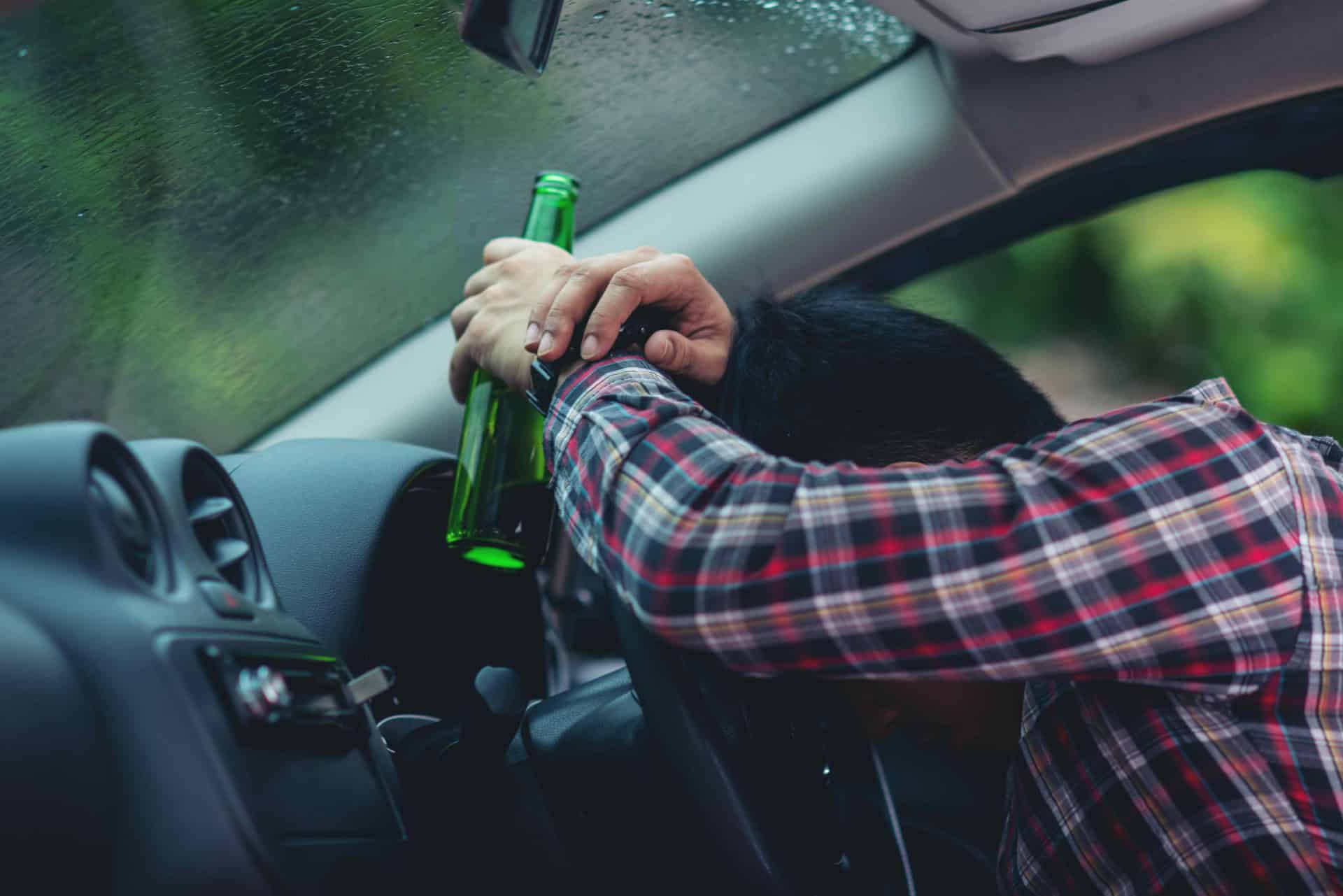 Person in a plaid shirt leans on a car steering wheel, holding a green bottle, with rain visible on the window.