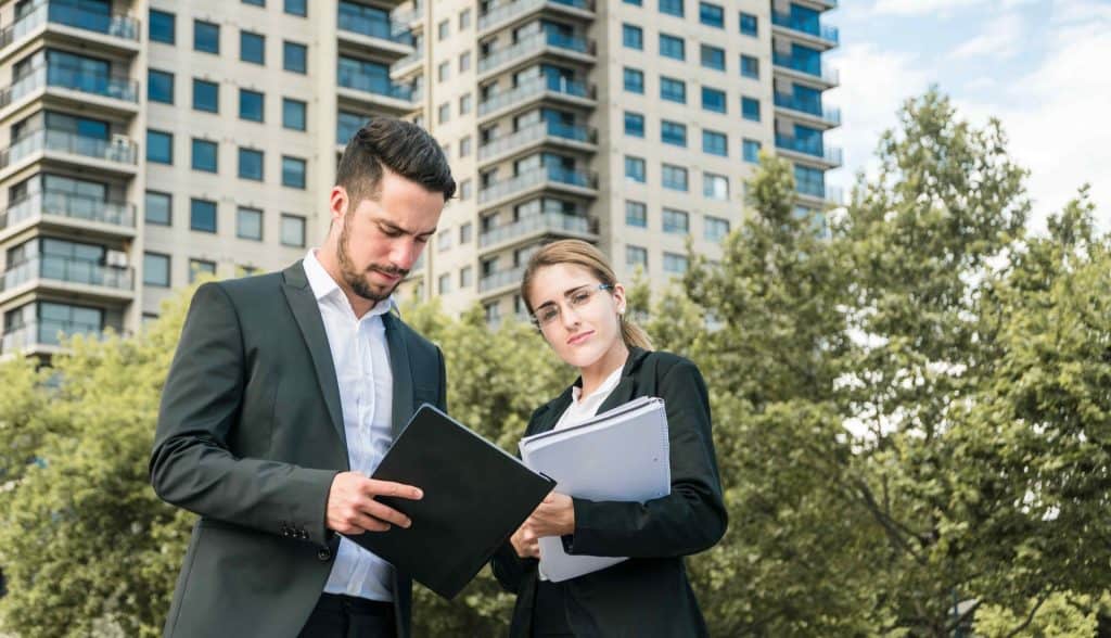 Two people in business attire stand outdoors, holding documents. A tall building and trees are in the background.