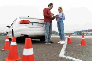 A man holding a clipboard talks to a woman near a white car on a driving course, with orange cones surrounding them.