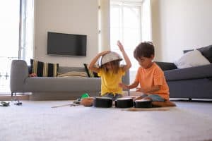 Two children play with pots and pans on a living room floor. One child holds a pot on their head, while the other taps on a pan. A sofa and TV are in the background.