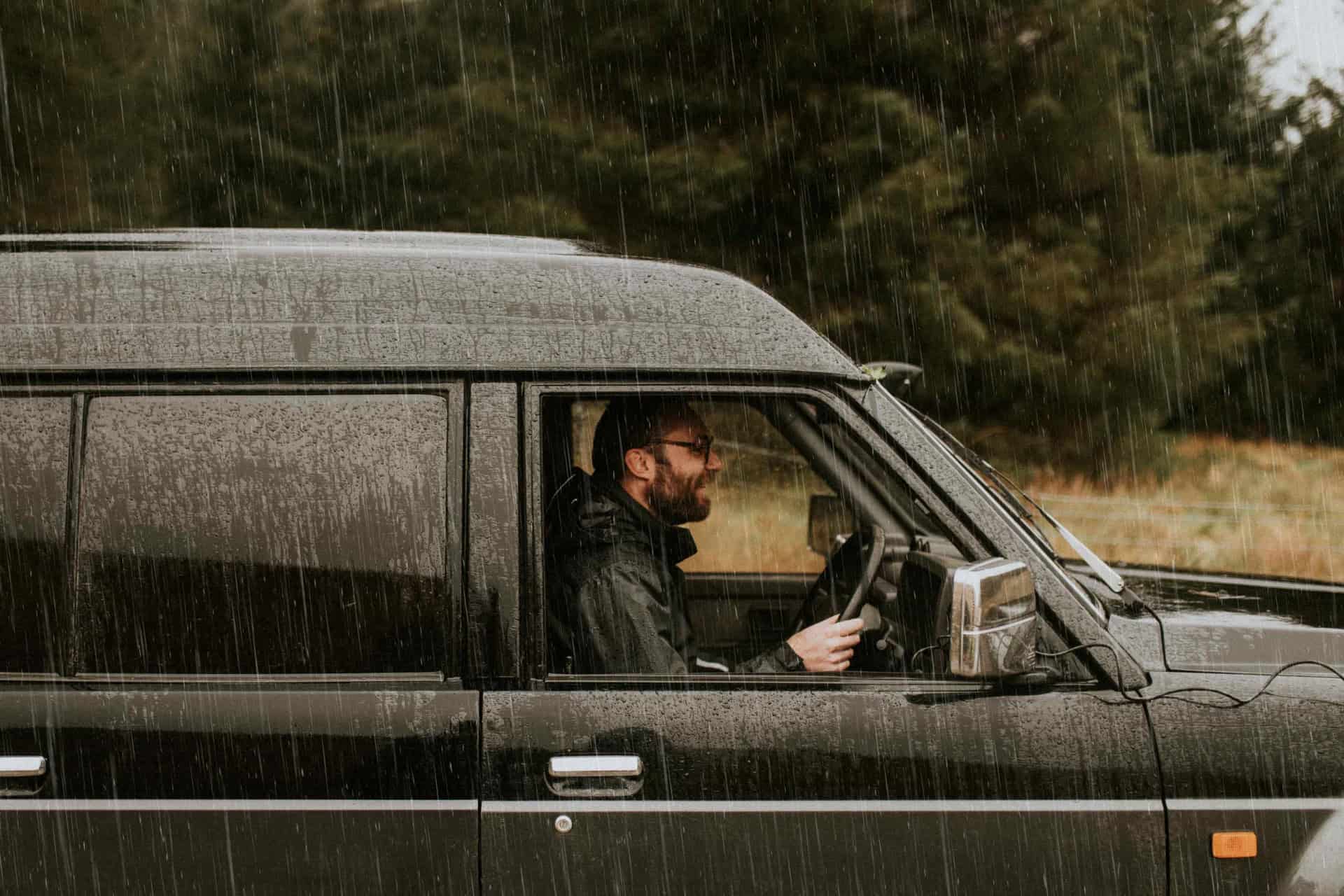 A bearded man drives a black vehicle through the rain, surrounded by greenery.