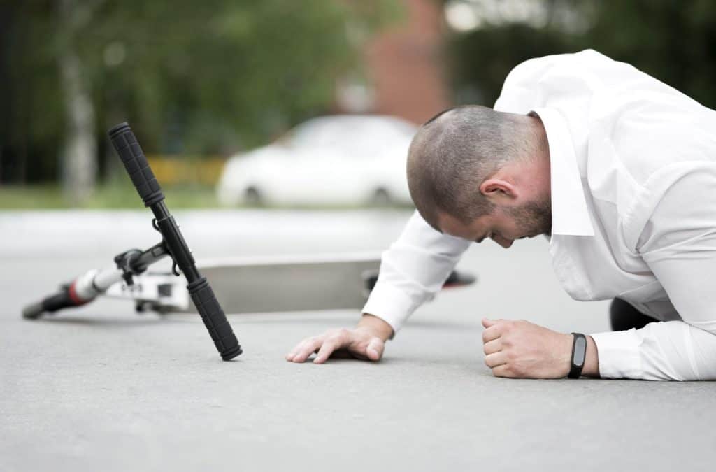 A man in a white shirt lies on the ground next to a fallen electric scooter on a concrete surface.
