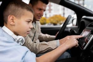 A child wearing headphones interacts with a touchscreen in a car, while an adult drives.