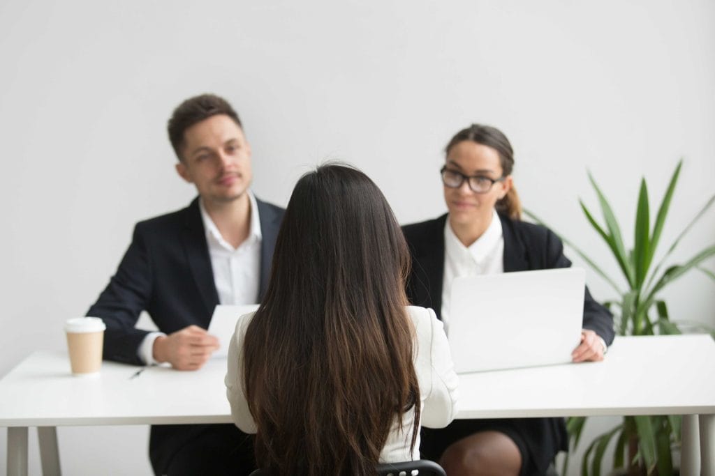 A woman sits across a table from two people in business attire, each with a laptop. A plant is in the background.