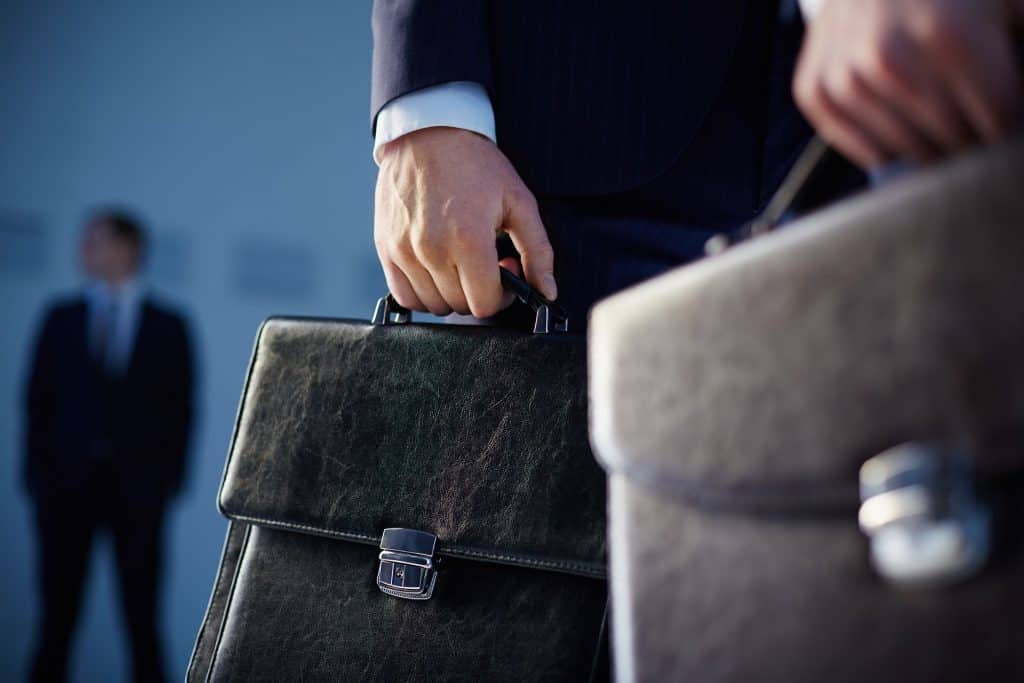Close-up of a person in a suit holding a black briefcase. Another person in a suit is blurred in the background.