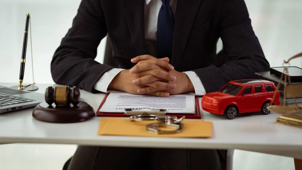Person in a suit sits at a desk with a laptop, gavel, legal documents, toy car, scales of justice, and a pen holder.