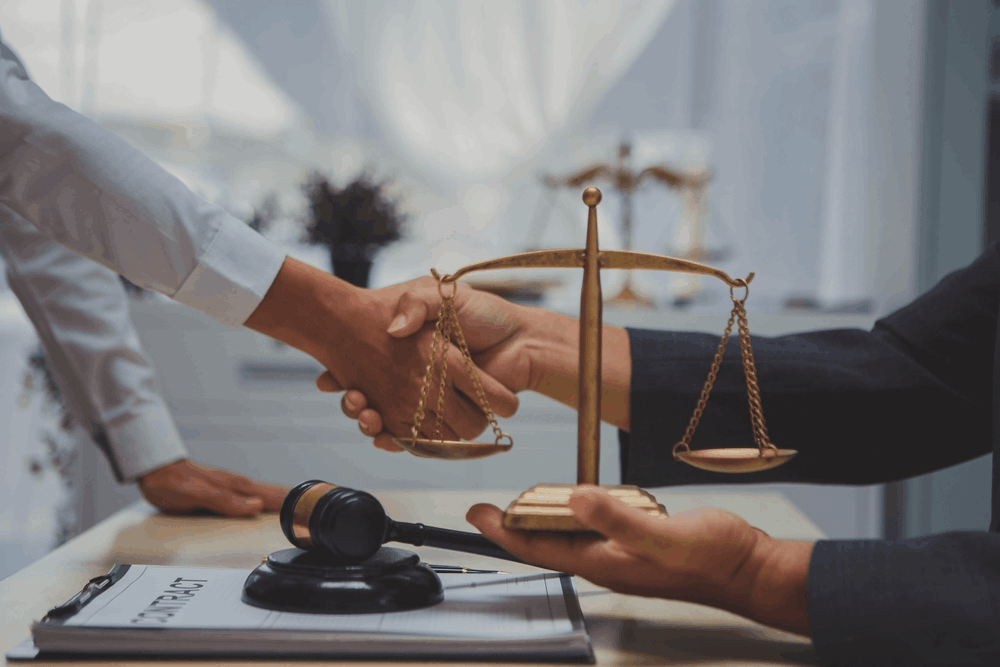 Two people shake hands over a wooden table with a gavel and scales of justice.