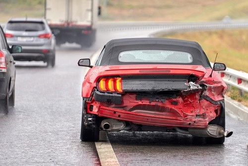 Red car with rear-end damage on a wet highway, surrounded by other vehicles.