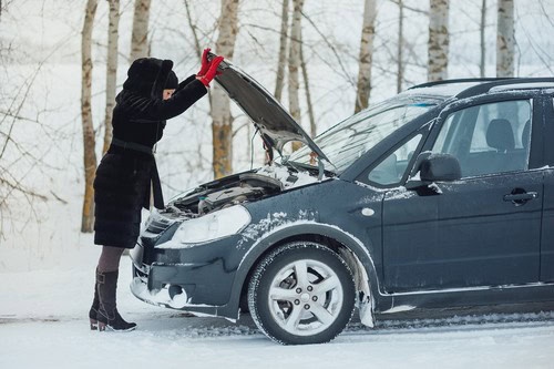 A person in winter clothing stands beside a snowy road, examining the engine of a car with the hood open.