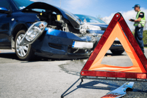 A damaged car with a crumpled hood sits on the road, next to a warning triangle in the foreground. In the background, a person in uniform stands watchfully, ready to assist. If you need guidance after such an incident, consulting an experienced attorney can provide clarity and support.