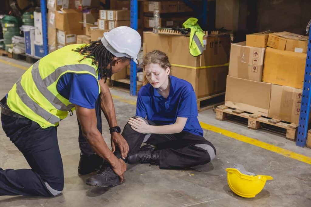 A worker in a safety vest kneels beside a colleague sitting on the floor holding their knee in a warehouse, with a hard hat lying nearby. If you need legal advice for workplace injuries, don't hesitate to contact our law office.