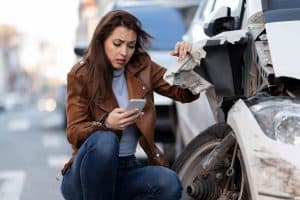 A woman kneels by a damaged car on the street, looking at her phone with concern. She's wearing a brown jacket and jeans.
