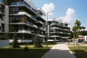 Modern multi-story apartment buildings with balconies, surrounded by green lawns and trees, under a partly cloudy sky. A paved pathway runs along the foreground.