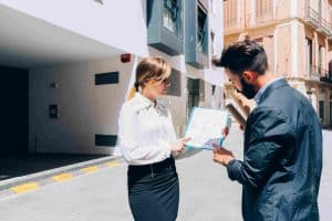 Two people stand outdoors discussing a blueprint. The woman points at the plan while the man holds it. They are near a modern building on a sunny day.