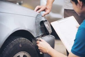 Person with a clipboard examines a dented car bumper. Another person points at the damage.