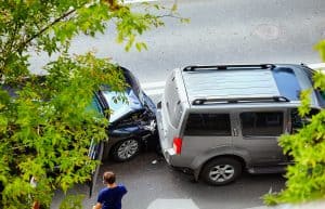 Two cars are involved in a rear-end collision on a street, surrounded by green leafy trees. A person in a blue shirt stands nearby.