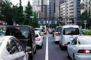Traffic in an urban area with multiple vehicles stopped at a red light, surrounded by tall buildings and trees.