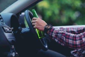 Person in a plaid shirt holding a green bottle while steering a car.