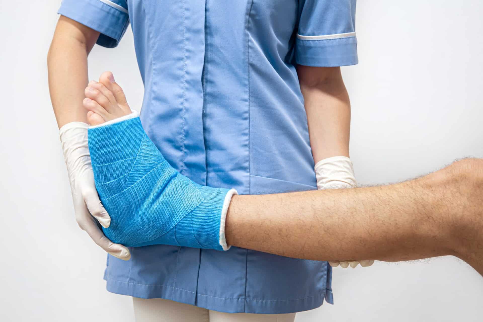 A person wearing a blue medical uniform holds the foot of another person, which is in a blue cast, against a plain white background.