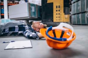 Worker lying on warehouse floor with eyes closed, next to a forklift. A hard hat and clipboard with pen are in the foreground.