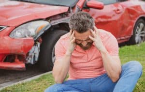 Man sitting on grass, holding his head in distress, with a damaged red car in the background.