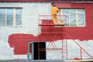 Worker on scaffolding painting a brick building wall red, with some areas still unpainted. Plastic covers the windows, and an open door is visible below.