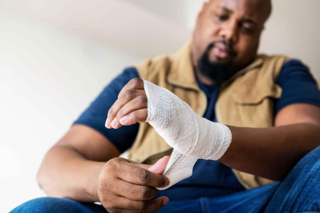 A man wraps a bandage around his hand while sitting down, wearing a blue shirt and beige vest.