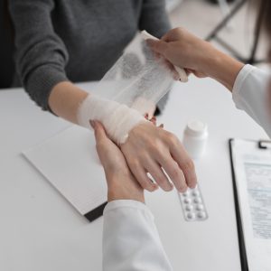 A medical professional wraps a bandage around a person's wrist at a desk with a clipboard, pills, and paper visible.