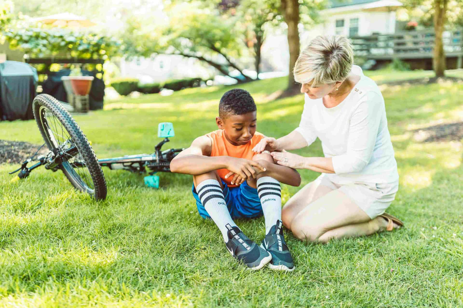 A woman tends to a boy's scraped knee after a minor bicycle accident, as he sits on the grass next to his fallen bike.