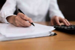 Person in a white shirt writing on paper with a pen while using a calculator on a wooden desk.