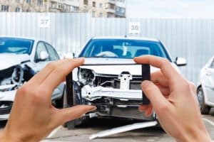 Person photographing the front damage of a white car using a smartphone, with another damaged car visible in the background.