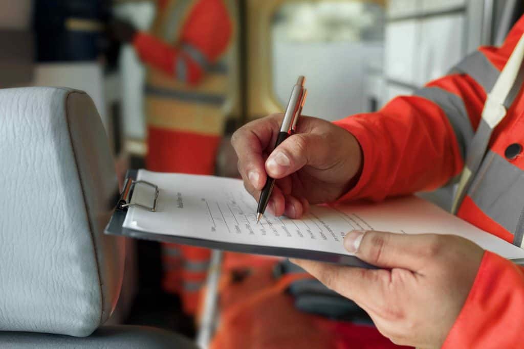 Person in a high-visibility orange jacket writing on a clipboard. Another person stands in the background, also wearing high-visibility clothing.