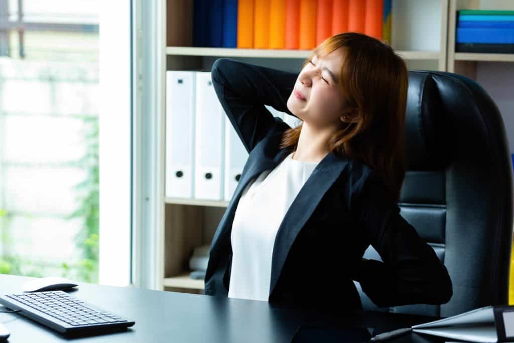 Person in a black blazer stretching and leaning back in an office chair near a desk, appearing to relieve tension.
