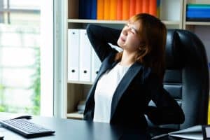 Person in a black blazer stretching and leaning back in an office chair near a desk, appearing to relieve tension.