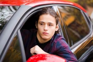 Person with long hair looks out from a red car's window, wearing a dark plaid shirt.