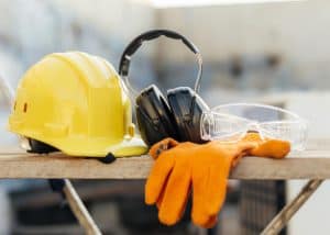 Yellow hard hat, black earmuffs, safety goggles, and orange gloves on a wooden plank.