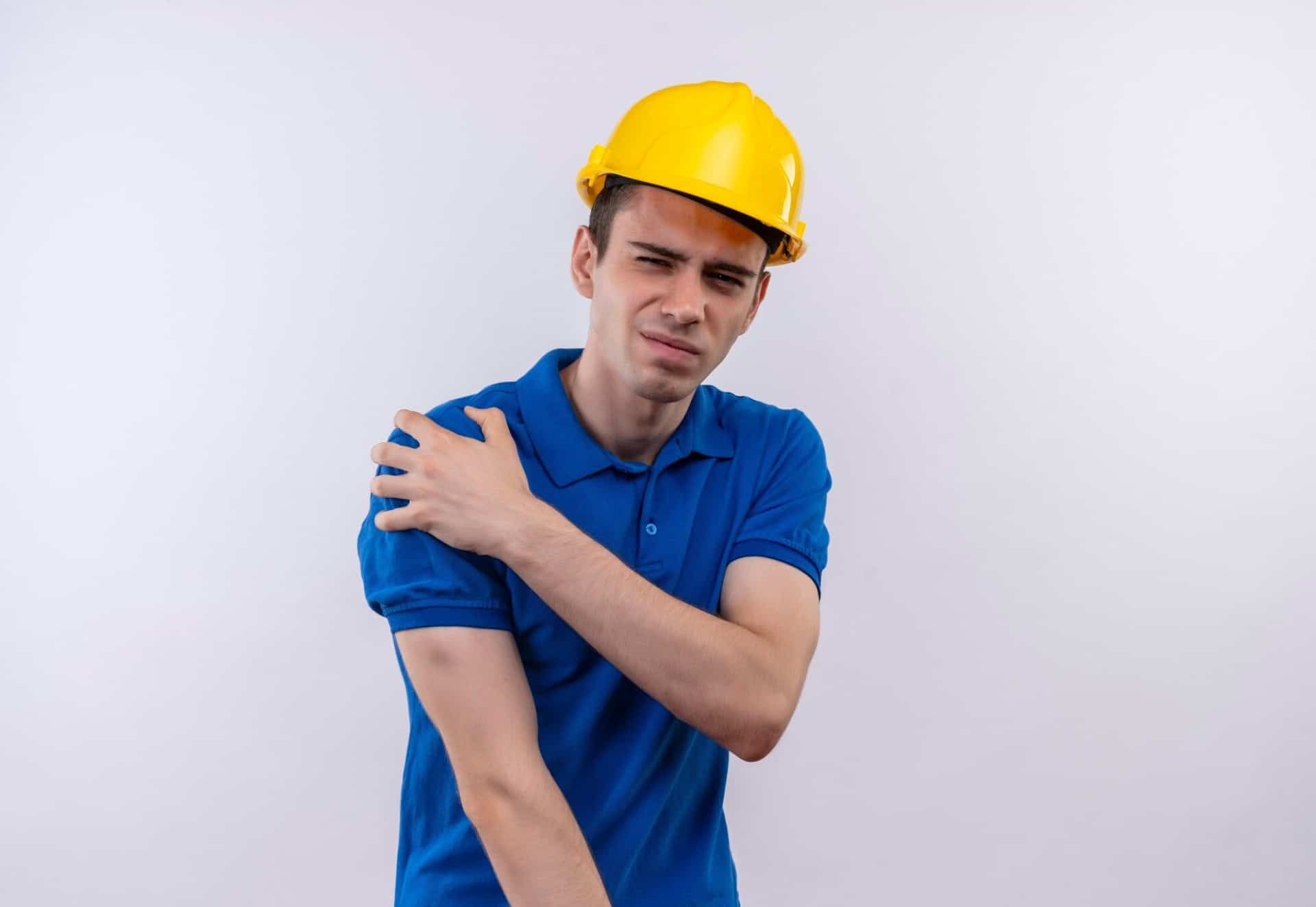 A person in a yellow hard hat and blue shirt stands with a pained expression, clutching their left shoulder, as if contemplating consulting an attorney for workplace injuries.