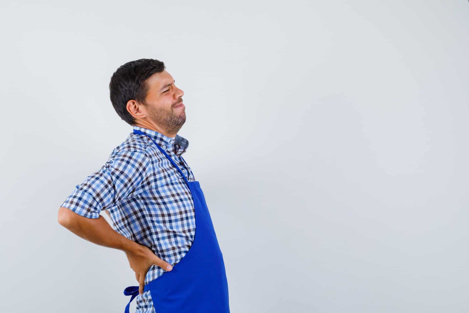A man in a blue apron and checkered shirt clutches his lower back with a pained expression, hinting at the physical toll of long hours at work.