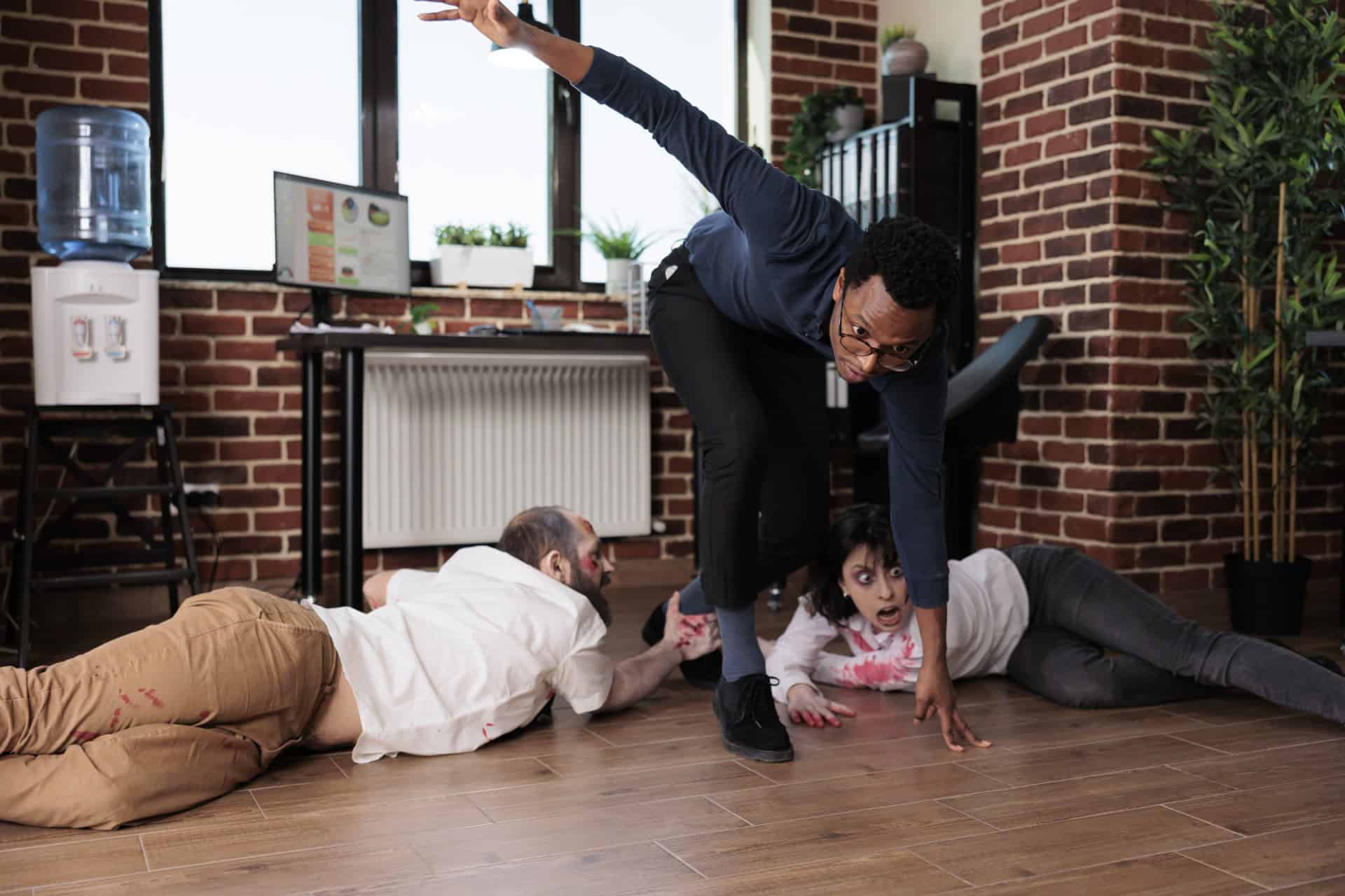 A person in business attire steps over two people on the floor in a brick-walled office, with one reaching out.
